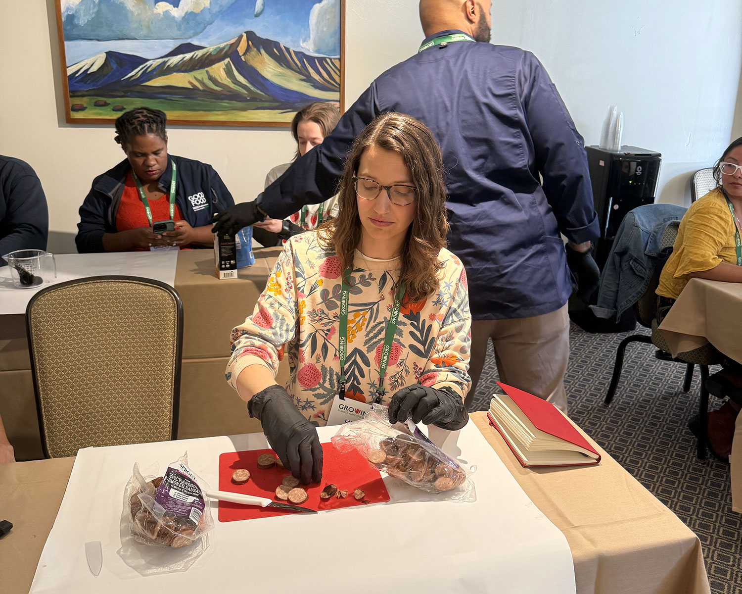 Woman wearing gloves slices sausage on a red cutting board during a hands-on food workshop, with other participants seated at tables in a conference room.