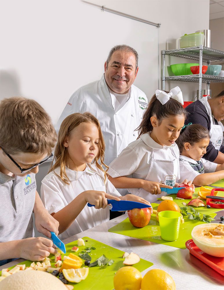 Chef stands behind elementary school students as they cut and prepare fresh fruit during a hands-on cooking activity.