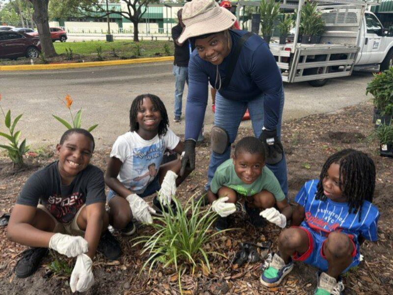 Adult volunteer and four elementary school students smile while planting and tending a garden bed outdoors.