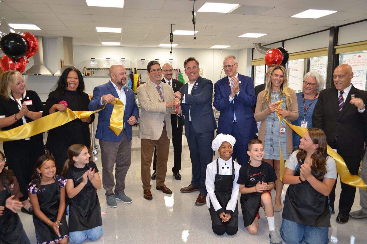 Students and adults celebrate a ribbon-cutting ceremony in a school culinary classroom, surrounded by gold ribbon and red and black balloons.