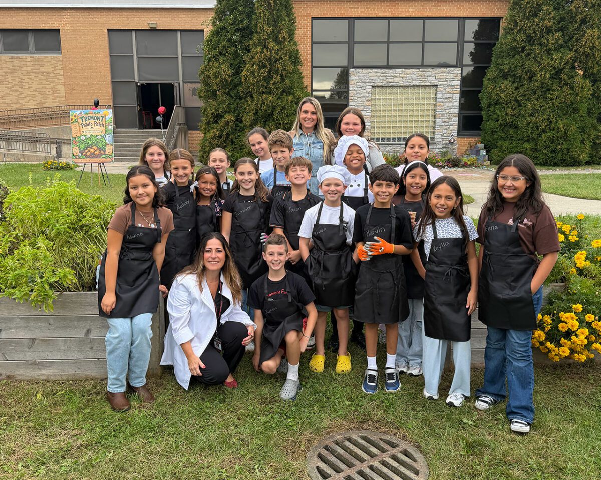 Students and teachers wearing black aprons pose together in the school garden beside raised vegetable beds and flowers.