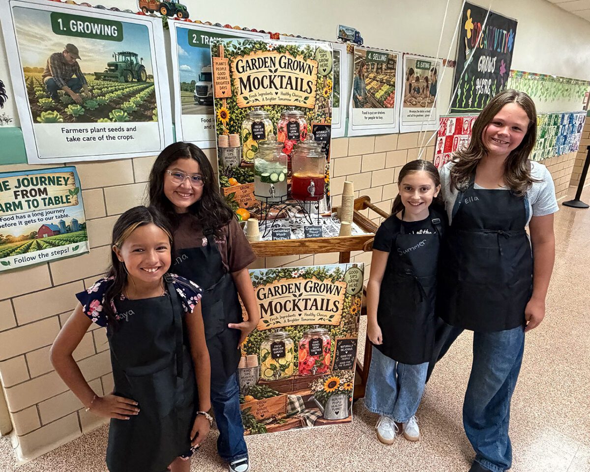 Four students in black aprons pose beside a Garden Grown Mocktails display featuring fresh, garden-inspired drinks and farm-to-table educational posters.